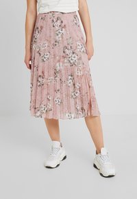 Kobieta w różowej plisowanej spódnicy midi w kwiatowy wzór z bocznym rozcięciem, w połączeniu z białymi sportowymi trampkami i białą bluzką w paski.