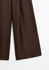 Hnědé široké kalhoty vyrobené z texturované látky, s pomačkaným povrchem a volným střihem. Žádný viditelný kovový prvek ani akcenty.