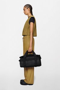 Borsa duffel nera con doppi manici, tessuto strutturato e design robusto, abbinata a un gilet kaki e pantaloni coordinati.