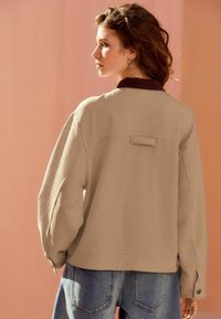 Veste beige texturée avec un col foncé, dotée d'un détail de patte au dos et de manches larges, assortie à un jean en denim bleu.