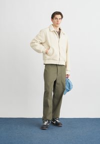Cream fleece bomber jacket with a collar, olive green wide-leg pants, and a light blue baseball cap. Black and white sneakers complete the outfit.