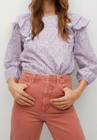 Helles lila Bluse mit floralen Mustern und Rüschendetails an den Schultern, kombiniert mit rosafarbenen cordartigen Hochbundhosen mit seitlichen Taschen.