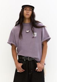 Fioletowy, oversize t-shirt z podwiniętymi rękawami, z małą grafiką na piersi. Połączony z czarnymi dżinsami i srebrnymi akcentami biżuteryjnymi.