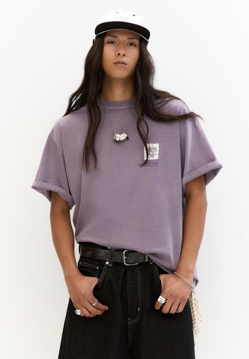 Paarse oversized t-shirt met opgerolde mouwen, met een kleine graphic op de borst. Gecombineerd met zwarte denim jeans en zilveren sierelementen.