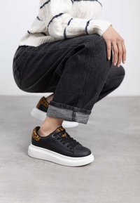 Zapatillas de cuero negro con suela blanca, detalles de estampado de leopardo en el talón y cordones negros. Combinadas con unos jeans oscuros con dobladillos.