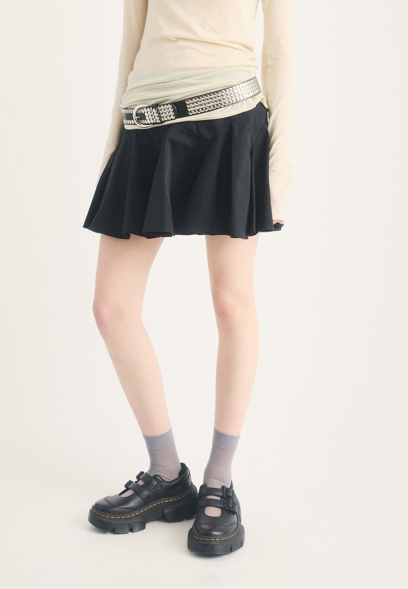 Personne portant une jupe évasée noire, un haut beige à manches longues, une ceinture cloutée, des chaussettes gris, et des chaussures noires à boucle, debout sur un fond uni.
