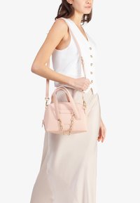 Piccola borsa a tracolla rosa realizzata in materiale testurizzato, con doppio manico, tracolla staccabile e dettaglio in catena dorata, esposta su uno sfondo bianco.