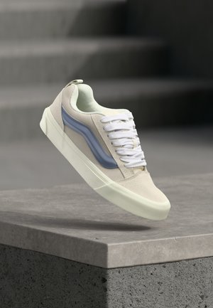 Pantof sport bej cu șireturi albe și dunga laterală albastră suspendat deasupra unei margini de beton, cu scări gri neclare în fundal.