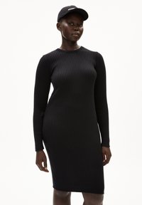 Schwarzes, geripptes Kleid mit figurbetonter Silhouette, langen Ärmeln und Rundhalsausschnitt. Dazu wird eine passende schwarze Mütze mit weißer Schrift getragen.