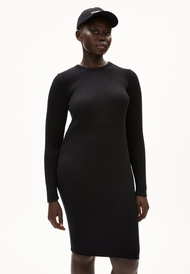 Schwarzes, geripptes Kleid mit figurbetonter Silhouette, langen Ärmeln und Rundhalsausschnitt. Dazu wird eine passende schwarze Mütze mit weißer Schrift getragen.