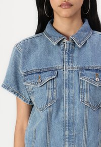 Camisa de mezclilla de manga corta, azul claro, con dos bolsillos en el pecho con herrajes plateados, detalles de botones y un diseño de cuello clásico.