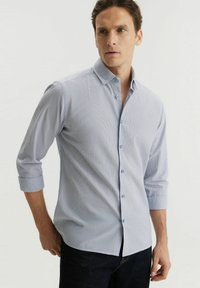 Jasnoniebieska koszula z guzikiem, z subtelnym wzorem w paski, długie rękawy podwinięte do łokci i klasyczny kołnierzyk. Tkanina wydaje się być fakturowana.