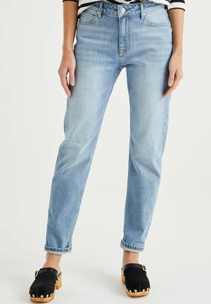 Lichtblauwe high waist jeans gedragen met zwarte houten klompen met gouden studs en een gestreept overhemd, getoond van taille tot voeten.