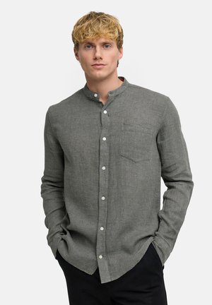 Junger Mann mit Händen in den Taschen, der ein strukturiertes graues Hemd mit Knopfleiste, Stehkragen und Brusttasche trägt, steht vor weißem Hintergrund.