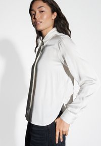 Camicia bianca a maniche lunghe con colletto, tessuto morbido, chiusura con bottoni e un leggero lucido. Indossata con jeans scuri, che mostrano un fit rilassato.