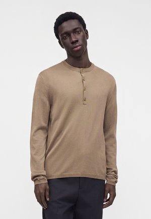 Joven con una camisa henley beige de manga larga con cuatro botones y pantalones oscuros, de pie frente a un fondo claro y liso.