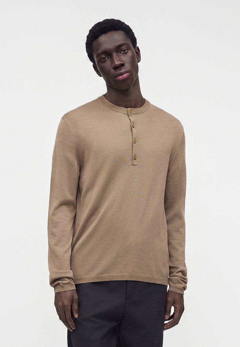 Jeune homme portant un t-shirt henley beige à manches longues avec quatre boutons et un pantalon foncé, se tenant devant un fond clair uni.