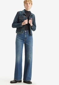 Spijkerjack met een vervaagde afwerking, voorzien van twee borstzakken en een kraag. Hoog-geplaatste, wijde blauwe jeans van stevige stof. Zwarte laarzen.