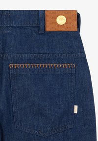 Des jeans en denim bleu foncé présentent une patch en cuir marron à la taille avec un logo embossé, des coutures orange sur la poche arrière et une étiquette blanche.
