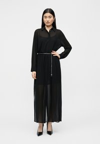 Μαύρο, διάφανο μακρύ φόρεμα πουκάμισο με γιακά, κουμπιά στο μπροστινό μέρος και μακριά μανίκια, σφιγμένο στη μέση με ασημένια αλυσίδα ζώνη.