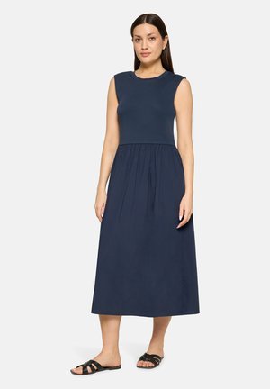 Vrouw staand in een mouwloze marineblauwe midi-jurk met een aansluitende top en een geplooide rok, gecombineerd met zwarte platte sandalen.