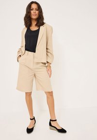 Beige linnblazer med två knappar, parat med matchande beige shorts. Svart linne under. Svarta kilklackar med ankelremmar.