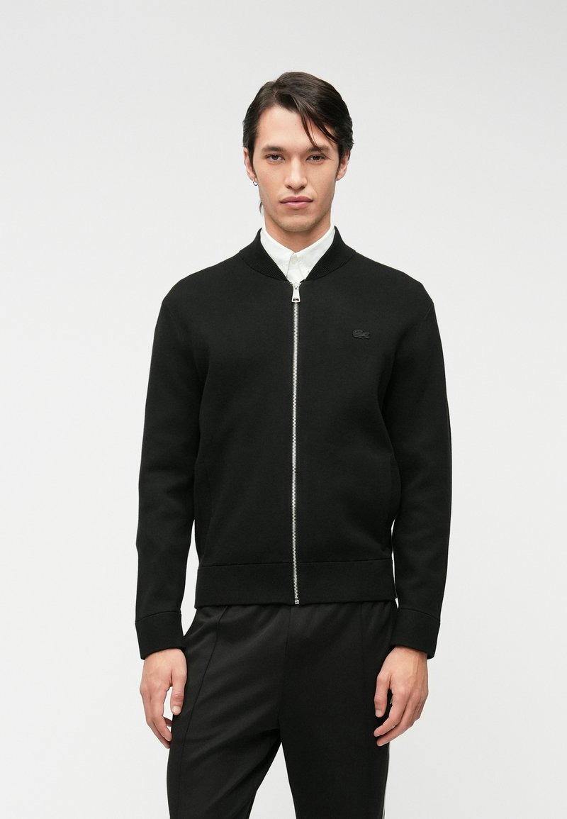 Μαύρο σακάκι bomber με φερμουάρ κατασκευασμένο από μαλακό ύφασμα. Διαθέτει ραμμένο γιακά και μανσέτες, με ένα μικρό λογότυπο στην αριστερή περιοχή του στήθους.