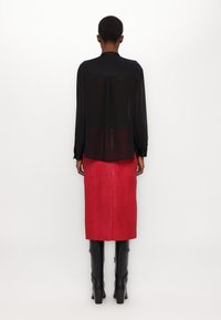 Blusa nera trasparente a maniche lunghe con colletto, abbinata a una gonna midi in pelle rossa uniforme e stivali neri al ginocchio con tacco.