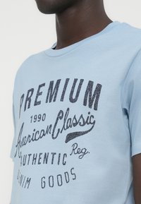 Κοντινό πλάνο ατόμου που φοράει γαλάζιο μπλουζάκι με μαύρο κείμενο που γράφει «Premium 1990 American Classic Authentic Reg. Denim Goods.»