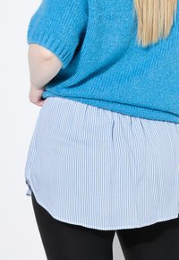Maglione blu a maglia con maniche corte indossato sopra una camicia bianca con sottili righe blu, abbinato a pantaloni neri.