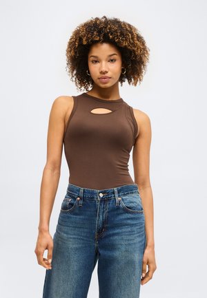 Γυναίκα με σγουρά μαλλιά που φοράει αμάνικη καφέ μπλούζα με άνοιγμα στο στήθος και ψηλόμεσο μπλε τζιν, στέκεται μπροστά σε απλό φόντο.