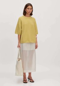 Helles gelbes, übergroßes Sweater, kurze Ärmel, mit einer strukturierten Oberfläche; kombiniert mit einem transparenten weißen Midikleid und schwarzen Flip-Flops, hält eine Stofftasche.