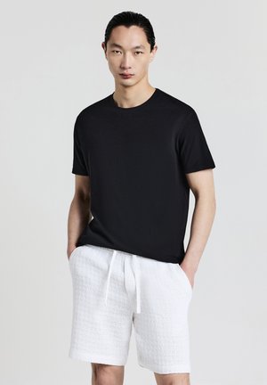 Mann mit dunklen Haaren, der ein schwarzes kurzärmliges Hemd und strukturierte weiße Shorts mit Kordelzug trägt, steht vor einem einfarbigen hellen Hintergrund.