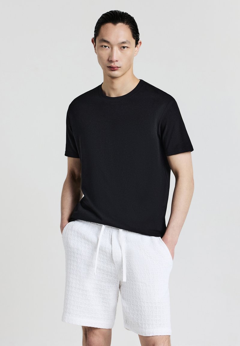Uomo con capelli scuri che indossa una camicia nera a maniche corte e pantaloni corti bianchi con coulisse texturizzati, in piedi contro uno sfondo chiaro e uniforme.
