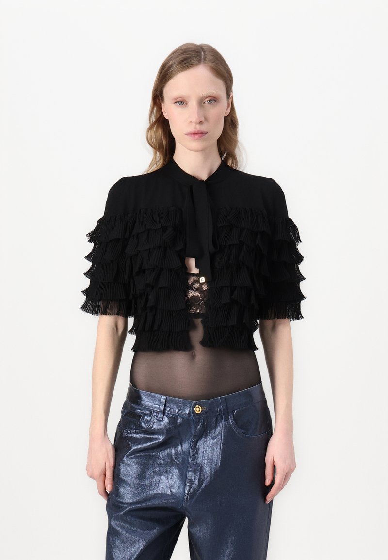 Top nero a maniche corte con dettagli in frange sovrapposte e chiusura a fiocco. Abbinato a pantaloni blu metallici con chiusura a bottone.