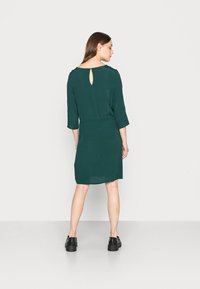 Vestido verde escuro, até ao joelho, com mangas três quartos e um detalhe em forma de buraco na parte de trás. Feito de um tecido leve e suave.