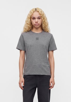 Joven con cabello largo, rizado y rubio que lleva una camiseta gris lisa de manga corta y pantalones oscuros, de pie frente a un fondo blanco.