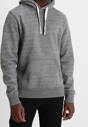 Personne portant un sweat à capuche gris uni avec des cordons blancs et une poche avant kangourou, assorti à un pantalon noir.