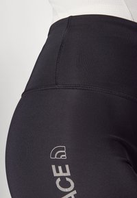 Černé legíny s vysokým pasem vyrobené z elastického materiálu, s hladkým povrchem a bílým logem potisknutým na straně.