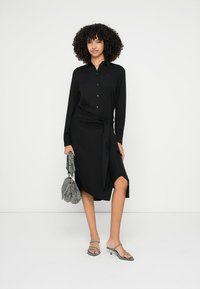 Crna haljina s dugim rukavima, remenom u struku, bočnim prorezom i kopčanjem na gumbe. Aksesoari uključuju teksturiranu sivu torbu i prozirne sandale.