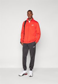 Rode zip-up jack, grijze joggingbroek en witte sneakers. Puma-logo's op alle items. De jack heeft een hoge kraag en elastische boorden.