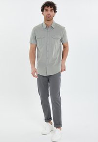 Camisa de botones de manga corta en gris claro con dos bolsillos en el pecho, combinada con pantalones oscuros ajustados y zapatillas blancas. Diseño y ajuste simples.