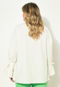 Witte blouse met uitlopende mouwen, vastgebonden bij de polsen. Glad, lichtgewicht stof; gestyled met groene broek. Achteraanzicht toont een vloeiende uitstraling.