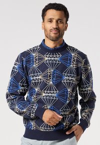 Man wearing navy blue sweater with geometric diamond-shaped patterns in blue, beige, and light blue, paired with light gray pants.
