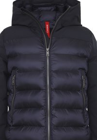 Giacca trapuntata nera con chiusura a zip, texture trapuntata e cappuccio. Presenta tasche laterali e un'etichetta rossa con logo sul colletto.