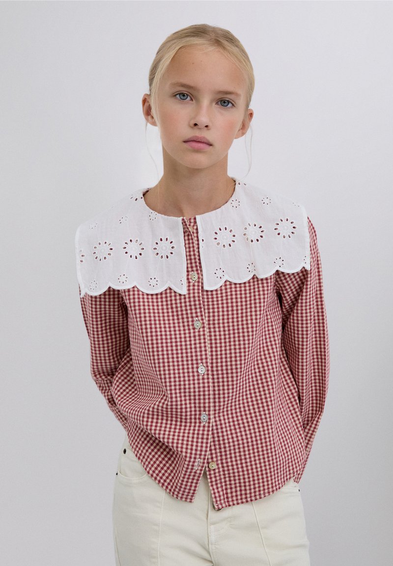 Red and white gingham shirt with a white eyelet lace collar, featuring a button front and long sleeves. Simple design and textured fabric.