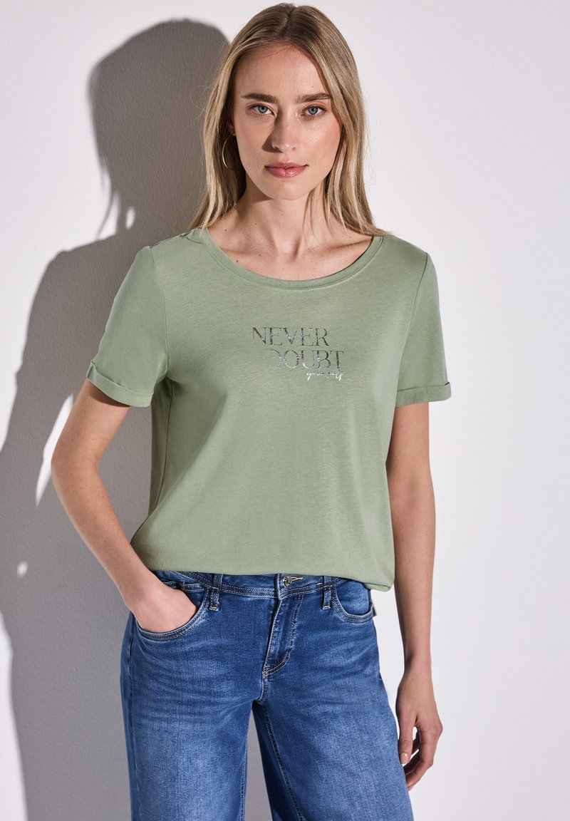 T-shirt vert clair à manches courtes avec un col rond, portant l'inscription "NEVER DOUBT" en lettres argentées. Associé à un jean en denim bleu.