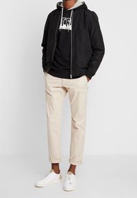 Chaqueta bomber negra sobre una sudadera gris, combinada con pantalones cremosos ajustados y zapatillas blancas con detalles en marrón. Tela suave, diseño casual.