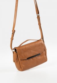 Braune Leder-Crossbody-Tasche mit Klappenverschluss, verstellbarem Riemen und einem subtilen schwarzen Akzent an der Vorderseite. Glatte Textur, rechteckige Form.