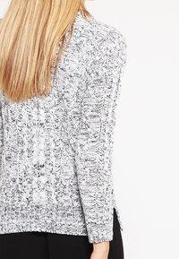 Strickpullover in Schwarz und Weiß mit hervorgehobenem Kabelmuster, langen Ärmeln und seitlichen Schlitzen am Saum für zusätzliche Designdetails.